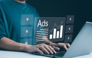 کەسێک لەسەر لاپتۆپ کار دەکات و چەندین ڤیدیۆ پلەیەری هۆلۆگرامی لە دەوروبەری دەبینرێن لەگەڵ گرافێکی "Ads" و چارتی ئاماری بۆ دیجیتاڵ مارکێتینگ