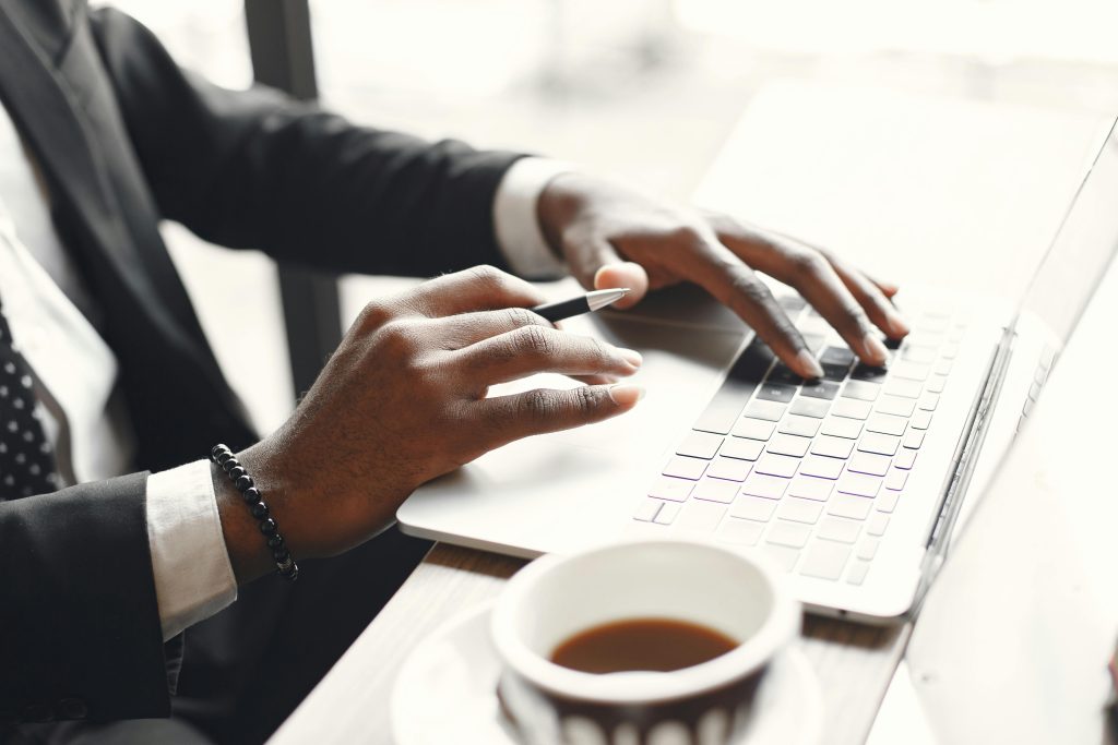 پیاوێک بە جلی کاری ڕەش و کراسی سپی لەسەر لاپتۆپێکی سپی کار دەکات، کوپێک قاوەی لە تەنیشتیەوەیە