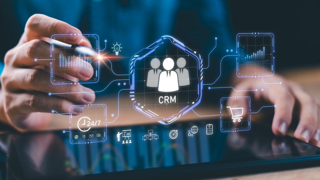 دوو کەس لە تابلێتێکدا کار دەکەن کە سیستەمی CRM یی دیجیتاڵی شینی لەسەر دەردەکەوێت، لەدەوریشدا ئایکۆنەکانی ئەنالیتیکس، پشتگیری 24/7، ئیمەیڵ، سەبەتەی کڕین، و ئامرازەکانی بازرگانی دیاری دەبن — وێنەیەکی کۆنسێپتی گۆڕانکاری دیجیتاڵی CRM.